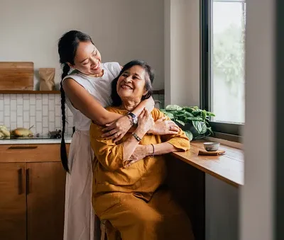 a happy beautiful woman hugging her mother while she is sitting in the kitchen and drinking